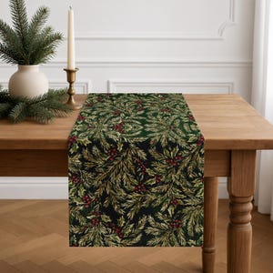 May include: A rectangular table runner featuring a dark green background with a repeating pattern of green pine branches, holly leaves, and small red berries. The fabric is draped over a wooden table surface, showcasing the detailed botanical print.