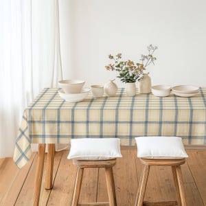 May include: A rectangular dining table covered with a cream and blue plaid tablecloth. On the table are several white ceramic bowls, a white ceramic vase with small flowers, and a white ceramic pitcher. Two wooden stools with white cushions.