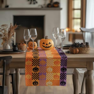 Può includere: Un runner da tavola rettangolare con un motivo ripetuto di quadrati a scacchi arancioni e viola intervallati da illustrazioni di zucche di Halloween arancioni e nere, esposto su un tavolo in legno con calici da vino.
