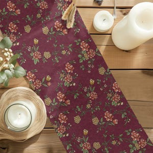 May include: A rectangular table runner featuring a deep burgundy background with a repeating floral pattern of red roses, small yellow flower clusters, and green vines. The fabric has a smooth texture and is laid out in a vertical orientation.