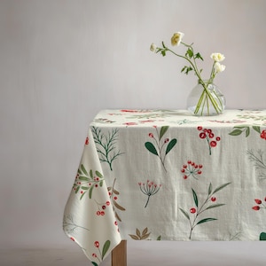 May include: A cream-coloured tablecloth featuring a repeating pattern of green botanical sprigs and small red berries. A clear glass vase sits on top of the tablecloth, containing three white flowers with green stems and leaves.