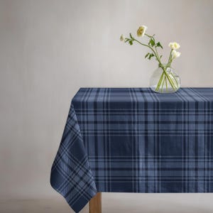 May include: A rectangular tablecloth featuring a dark blue and light blue plaid pattern with intersecting horizontal and vertical lines. A clear glass vase sits on the surface, containing three white flowers with green stems and leaves.