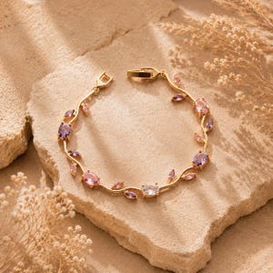 Può includere: Un bracciale color oro caratterizzato da una delicata catena a forma di vite, decorata con pietre preziose sfaccettate di forma ovale nei colori rosa, viola e trasparente. Il gioiello presenta una chiusura sicura e piccoli dettagli a foglia.