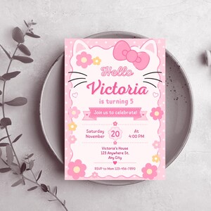 A pink cat-themed birthday invitation with a large pink bow, cat ears, and whiskers, resting on a gray ceramic plate with green leaves in the background.