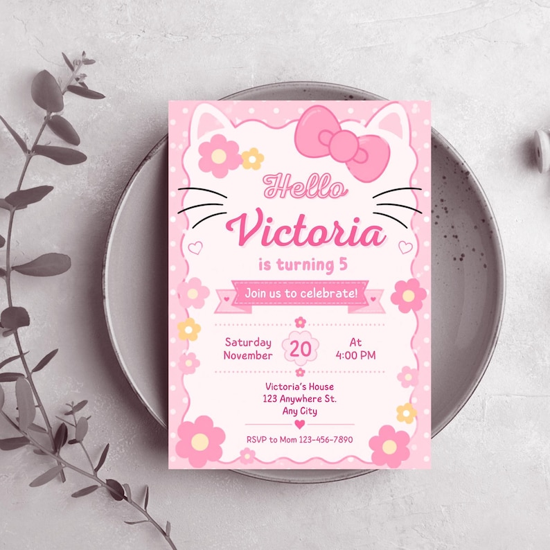 A pink cat-themed birthday invitation with a large pink bow, cat ears, and whiskers, resting on a gray ceramic plate with green leaves in the background.