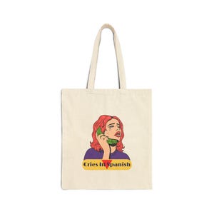 May include: A beige canvas tote bag featuring a graphic illustration of a person with red hair crying while holding a green telephone receiver to their ear. Below the illustration, the text Cries In Spanish is printed in a yellow oval.