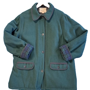 May include: A dark green wool-blend jacket featuring a button-front closure, a pointed collar, and two front flap pockets with dark trim. The sleeve cuffs are turned up to reveal a blue and green plaid lining. A Woolrich label is at the neck.