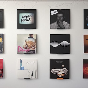 May include: A collection of twelve square vinyl record covers displayed in a grid on a wall. The covers feature various musical artists and album titles including Daft Punk, John Mayer, La La Land, and Dawn FM, held by small black and orange wall mounts.