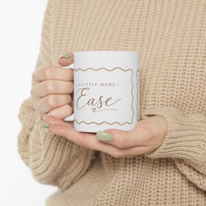 Puede incluir: Una taza de cerámica blanca con un marco dorado de bordes ondulados y el texto A LITTLE MORE Ease junto a un pequeño icono de corazón en el centro. La taza tiene un acabado liso y un asa estándar con un diseño limpio y sencillo.