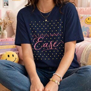 Puede incluir: Una camiseta azul marino de manga corta con un estampado de pequeños corazones amarillos y el texto A LITTLE MORE Ease impreso en rosa, en letra cursiva y de imprenta en la parte delantera. La camiseta se combina con vaqueros azules y dos pulseras.
