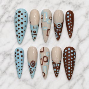 May include: A set of ten almond-shaped press-on nails featuring a variety of designs in shades of light blue, brown, and beige. The patterns include brown polka dots on light blue, geometric squares, and circular motifs on neutral backgrounds.