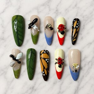 May include: A set of ten almond-shaped press-on nails featuring various designs including green leaf patterns, butterfly wing motifs, and three-dimensional insect embellishments such as ladybugs and bees, painted in vibrant colors like red, blue, and yellow.
