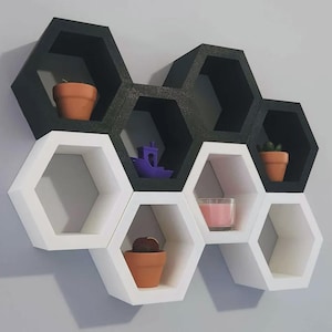May include: A wall-mounted shelving unit composed of seven interconnected hexagonal compartments. The top three shelves are black, and the bottom four are white. The shelves contain three terracotta pots, a small purple boat, and a pink candle.