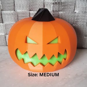 May include: A medium-sized, orange, geometric pumpkin decoration with a black stem and a carved, jagged face revealing a bright green interior. The pumpkin features sharp, angular facets and a menacing expression with triangular eyes and a wide, toothy grin.