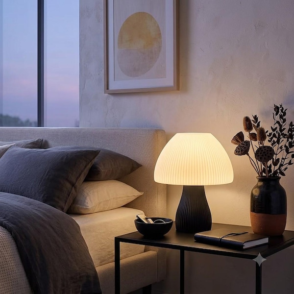 May include: A mushroom-shaped table lamp with a ribbed white shade and a black base, positioned on a black metal side table next to a black ceramic vase containing dried botanicals and a closed black notebook.