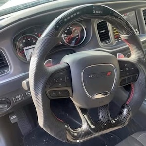 May include: A black steering wheel with a carbon fibre top and bottom, perforated leather side grips, and red stitching. The centre features a Dodge emblem, integrated control buttons, and paddle shifters, positioned in front of a vehicle dashboard.