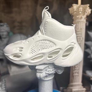 May include: A white high-top sneaker featuring a textured, perforated side panel and a thick, sculpted sole with multiple oval cutouts. The shoe has a smooth upper with a pull tab at the heel and a series of eyelets along the lace-up area.