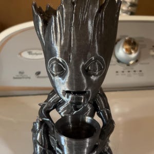 May include: A dark gray, metallic-finish planter shaped like a humanoid tree character with large eyes and a textured, bark-like surface. The figure is depicted in a seated position, holding a small, cylindrical pot in its hands, with an open top.