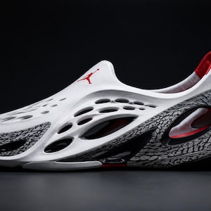 May include: A white slip-on shoe featuring a perforated design, a red Jumpman logo on the upper, and a grey and black elephant print pattern along the sides and heel. The interior lining is red, and the sole has black and red accents.