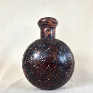 May include: A spherical vase with a narrow neck and a rounded rim. The surface is dark brown with a distressed, textured finish and features a repeating pattern of small, light-colored, upward-facing floral motifs etched across the body.