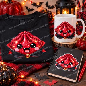 Może przedstawiać: Czarny t-shirt, biały ceramiczny kubek i czarny notatnik spiralny, każdy z grafiką brokatowego czerwonego pierożka w czarnej pelerynie wampira ze złotą broszką, dużymi czarnymi oczami i dwoma małymi białymi kłami.