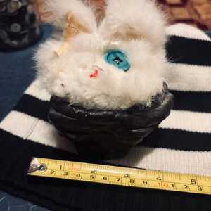 May include: A white fuzzy plush creature with a single blue button eye and a small red stitched mouth, seated in a dark textured bowl. A yellow measuring tape is positioned in front, showing a width of approximately 5.25 inches.