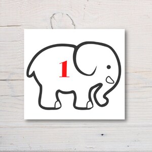 May include: A white square with a black outline drawing of an elephant. The elephant has a red number 1 in the middle. The image is on a white wooden surface.