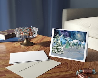 May include: A greeting card with a winter landscape scene featuring snow-covered trees and mountains. The card is on a wooden table with a white envelope and a clear glass dish filled with candies. A book and a brass-footed dish are also on the table.