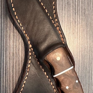 May include: A fixed-blade knife with a polished brown burl wood handle featuring three brass pins and a white spacer, partially inserted into a dark brown leather sheath with thick, light-coloured stitching and two integrated belt loops.