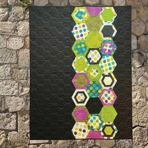 May include: A handmade quilt with a black background and a vertical column of colorful hexagon shapes. The hexagons feature various patterns, including polka dots and geometric designs, in shades of green, pink, and blue. The quilt is displayed against a stone wall.