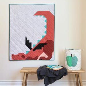 May include: A quilted wall hanging featuring a red dragon design with turquoise accents and a teal border. The dragon is set against a white background with a diamond pattern. A wooden bench holds a dark gray blanket, books, and a white storage bin with a green apple graphic.