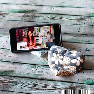 May include: A cell phone stand with a floral pattern, supporting a smartphone displaying a video. The stand is made of fabric with a gray background and white and gold flowers. The text "CELL PHONE STAND" is at the top.