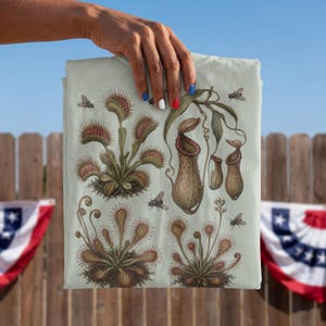 May include: A light-colored t-shirt featuring a detailed botanical illustration of various carnivorous plants, including Venus flytraps, pitcher plants, and sundews, interspersed with small flying insects against a pale background.