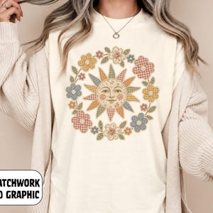 Peut inclure: Un t-shirt crème arborant un motif central représentant un soleil à visage humain, entouré d'une couronne de fleurs. Le soleil et les fleurs présentent un motif faux patchwork dans des tons sourds de bleu, jaune, rose et vert.