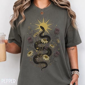 Puede incluir: Una camiseta gris carbón con un estampado gráfico de una serpiente negra enroscada entre margaritas amarillas y cardos morados, bajo un sol amarillo estilizado con líneas radiantes. El diseño presenta detalles finos y elementos botánicos.