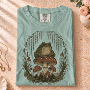 Op de afbeelding: Een licht saliegroen t-shirt met een grafische illustratie van een pad die op een grote paddenstoel zit, omringd door wilgentakken en lisdodden. Het label in de nek toont de tekst Comfort Colors en de maat M.