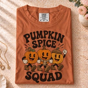 Peut inclure: Un t-shirt couleur rouille avec un motif de trois citrouilles de dessin animé dotées de visages, de bras et de jambes. Le texte au-dessus indique PUMPKIN SPICE et en dessous SQUAD. L'étiquette Comfort Colors à l'intérieur indique la taille M.