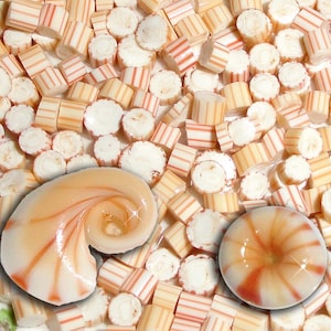 May include: A close-up of a pile of candy canes with red and white stripes. There are two seashells in the image, one is a spiral shape and the other is a round shape. Both seashells are a light orange color with red and white stripes.