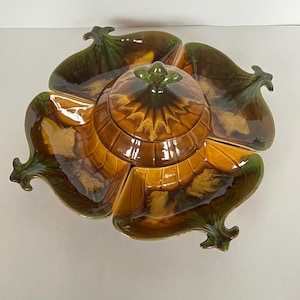 May include: A ceramic serving dish featuring a central lidded bowl surrounded by four leaf-shaped compartments. The piece has a glossy glaze in shades of amber, brown, and deep green, with a textured finish and a small knob on the lid.