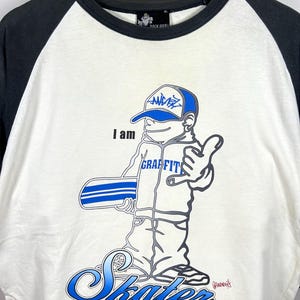 May include: A white and black raglan t-shirt featuring a graphic of a cartoon character wearing a cap and holding a skateboard. The text reads "I am GRAFFITI Skater". The design uses blue, black, and white colors, with a round neckline and short sleeves.