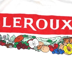 以下が含まれることがあります： 白い布地に、赤い長方形のグラフィックと白い文字で「LEROUX」と書かれています。文字の周りには、ブラックベリー、イチゴ、オレンジ、リンゴ、サクランボ、ブルーベリー、バナナなど、さまざまな果物のカラフルなイラストが描かれています。