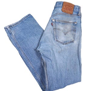 May include: Light blue denim jeans featuring a faded wash, a tan leather patch on the back waistband, and a small red tab on the back pocket. The jeans have a classic five-pocket design with visible stitching and a button fly closure.