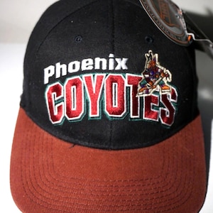 Peut inclure: Une casquette de baseball noire avec une visière marron, ornée du texte brodé Phoenix Coyotes en blanc et rouge avec un contour vert. Un petit logo coyote brodé est placé à droite du texte. Une étiquette de produit officiel NHL est attachée.