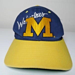 May include: A navy blue baseball cap featuring a large, embroidered yellow letter M in the centre. The word Wolverines is stitched in white script across the upper portion of the crown. The cap has a solid yellow brim and small yellow eyelets.