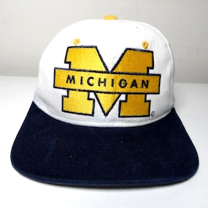 May include: A white baseball cap with a dark blue brim. The front features a large, embroidered yellow block letter M with the word MICHIGAN stitched in dark blue across the centre. A small yellow button sits at the top centre of the crown.