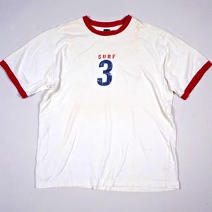 May include: A white short-sleeved ringer t-shirt with red trim on the crew neckline and sleeve cuffs. The centre of the chest features the red text "SURF" positioned above a large, distressed blue number "3" graphic print.