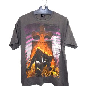 May include: A charcoal gray short-sleeve t-shirt featuring a graphic print of a tombstone with a cross on top. The text on the tombstone reads "HERE LIES SPIDER-MAN". A dark, shadowy figure is kneeling in front of the tombstone against a background of pink diagonal stripes and lightning.