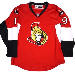 May include: A red, white, and black Reebok hockey jersey featuring the Ottawa Senators logo on the chest. The jersey has a V-neck collar, the number 19 on the sleeves, and an NHL shield logo centered below the collar. The design includes color-blocked sleeves.