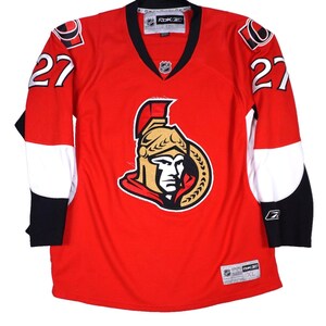 May include: A red Reebok hockey jersey featuring the Ottawa Senators logo on the chest, the number 27 on both sleeves, and black and white accents. The collar is black with a V-neck design, and the jersey includes NHL and Reebok branding tags.