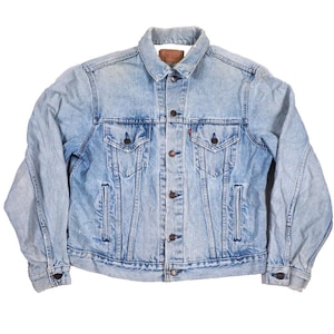 Könnte beinhalten: Eine hellblaue Jeansjacke mit klassischem Kragen, einer durchgehenden Knopfleiste mit Metallknöpfen, zwei Brusttaschen mit geknöpften Klappen, zwei seitlichen Eingrifftaschen sowie langen Ärmeln mit geknöpften Manschetten und einem Lederetikett.